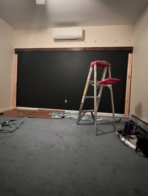 Bedroom mid-renovation with a freshly painted dark wall, bare floor, drop cloths, and a step ladder